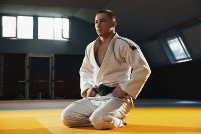 Young judo caucasian fighter in blue kimono with black belt posing confident in the gym, strong and healthy. Practicing martial arts fighting skills. Overcoming, reaching target, self building up.